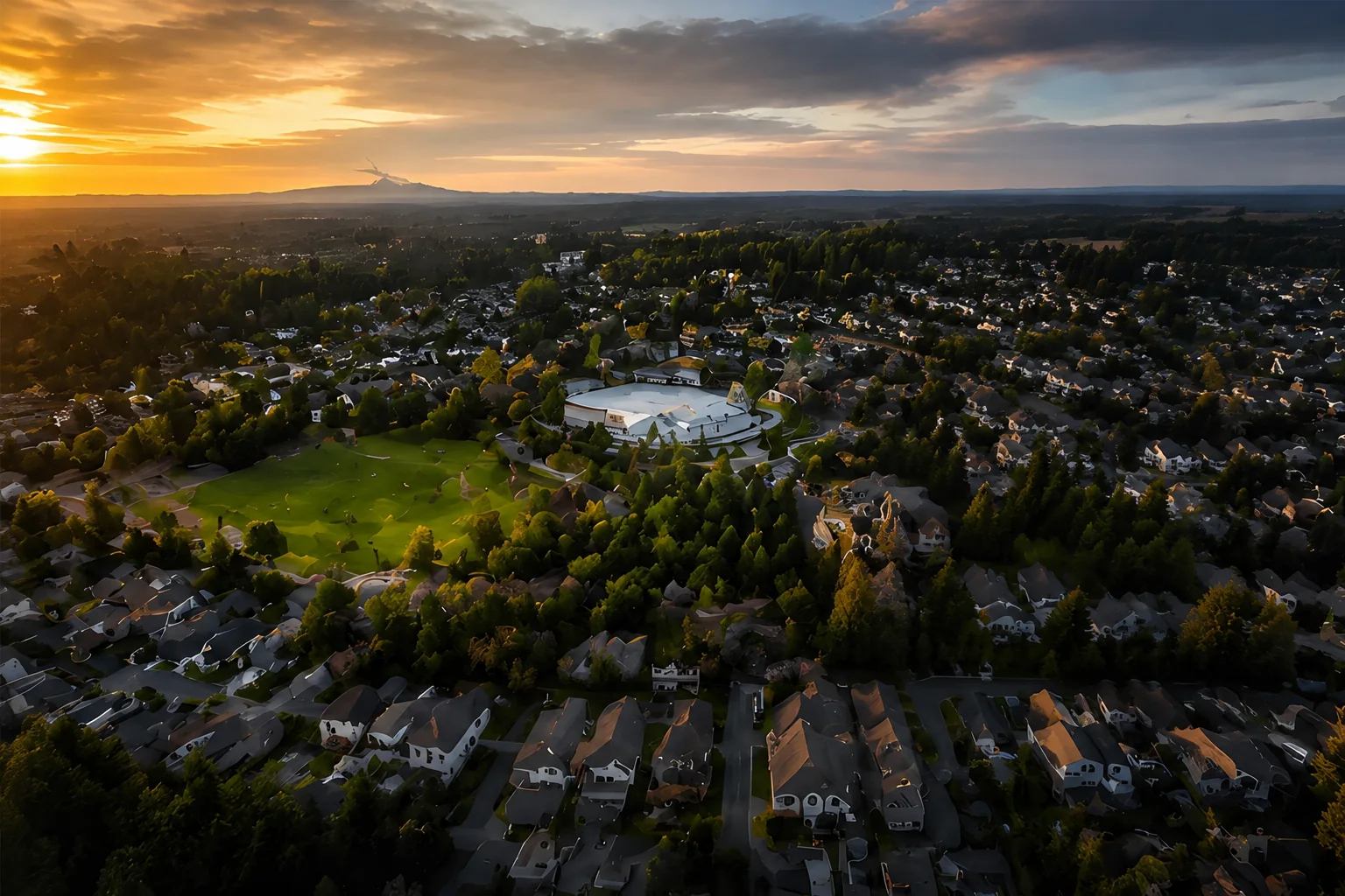 Golden hour aerial over Langley BC showing tree lined residential streets, rooftops, a central green playing field and low hills on the horizon