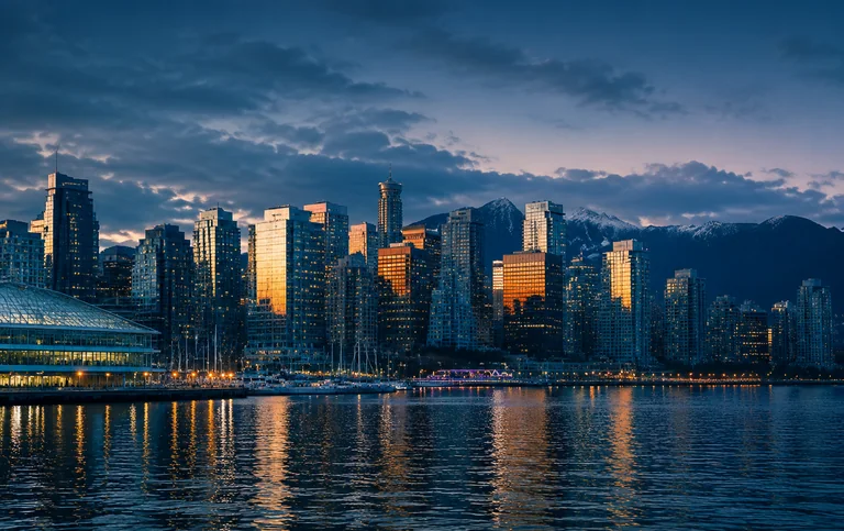 A downtown waterfront skyline seen across the harbour at dusk, with the North Shore mountains behind