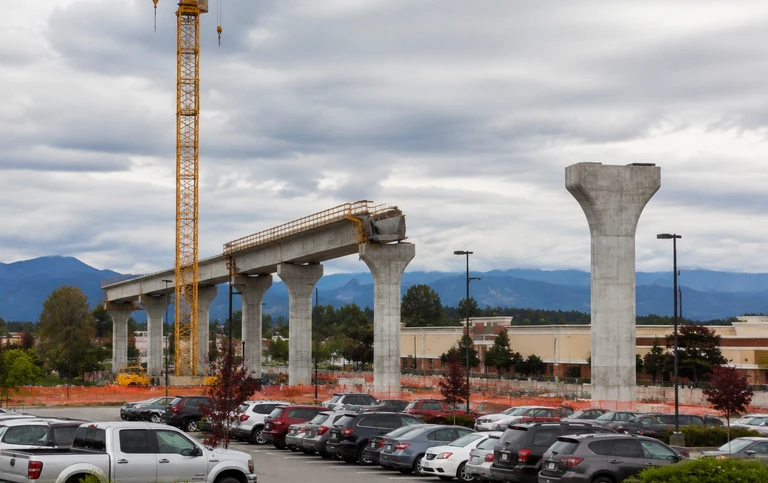 An elevated transit guideway under construction beside a suburban shopping centre car park, with bare concrete piers and a crane and no track laid yet