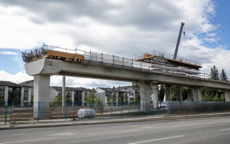 An elevated transit station under construction on a suburban arterial road, with raw concrete piers, a half finished platform deck and construction hoarding
